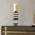 A modern table candle holder with a black and clear striped acrylic column, a rose gold cone-shaped accent, and a white pillar candle on top, displayed on a black console table against a textured gray wall.
