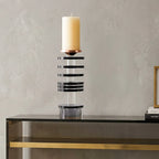 A modern table candle holder with a black and clear striped acrylic column, a rose gold cone-shaped accent, and a white pillar candle on top, displayed on a black console table against a textured gray wall.