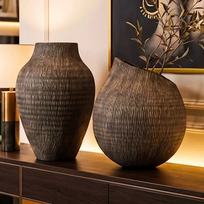 A styled interior scene with a dark wood console table holding two large textured vases, beneath a framed portrait artwork on a beige wall. The room includes a green velvet chair, fireplace, shelves, and warm ambient lighting.