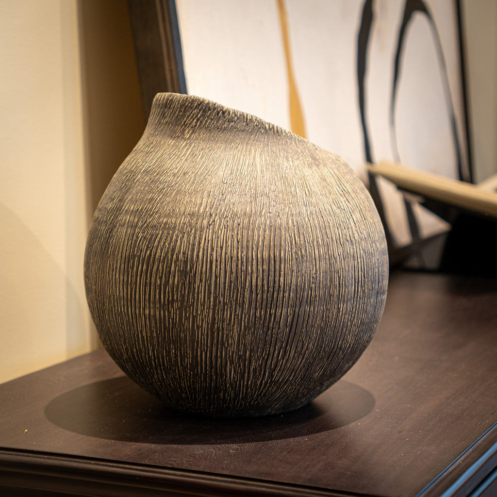 A textured, rounded ceramic vase with a unique asymmetrical spout, displayed on a dark wooden table in an indoor setting.