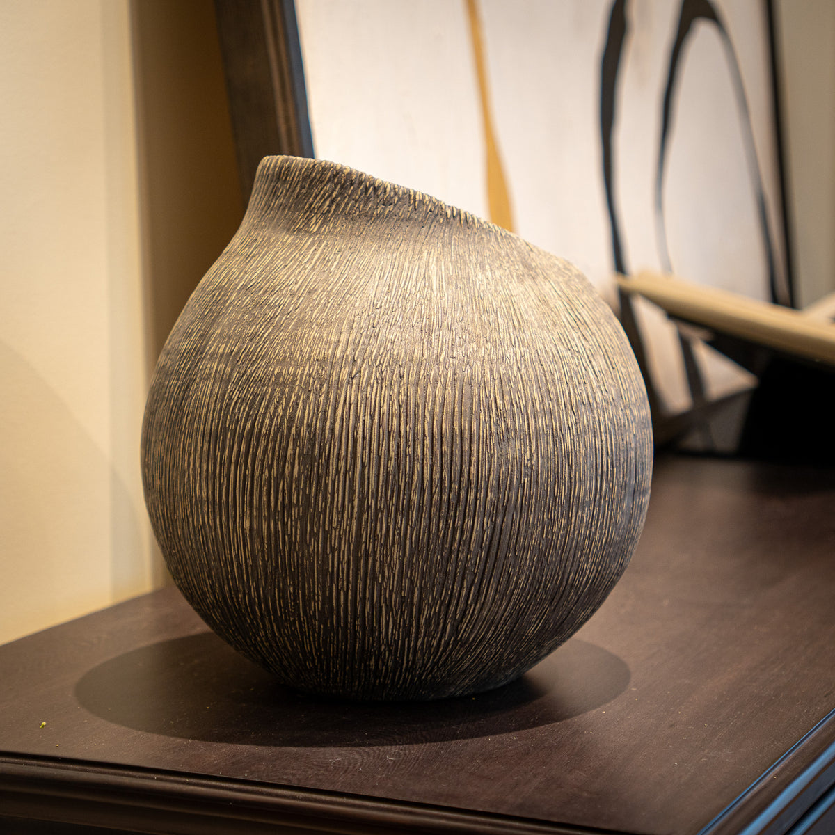 A textured, rounded ceramic vase with a unique asymmetrical spout, displayed on a dark wooden table in an indoor setting.