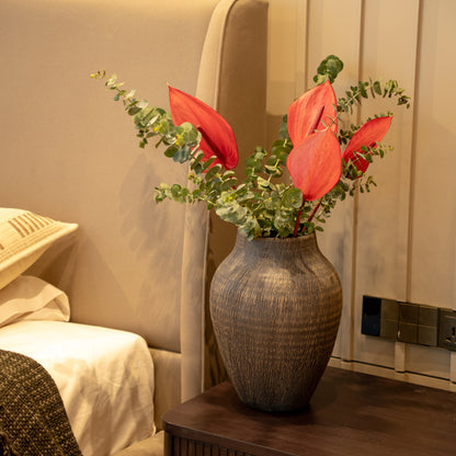 A textured dark brown ceramic vase filled with red anthuriums and green eucalyptus stems, styled on a bedside table next to a bed in a warm-toned bedroom.