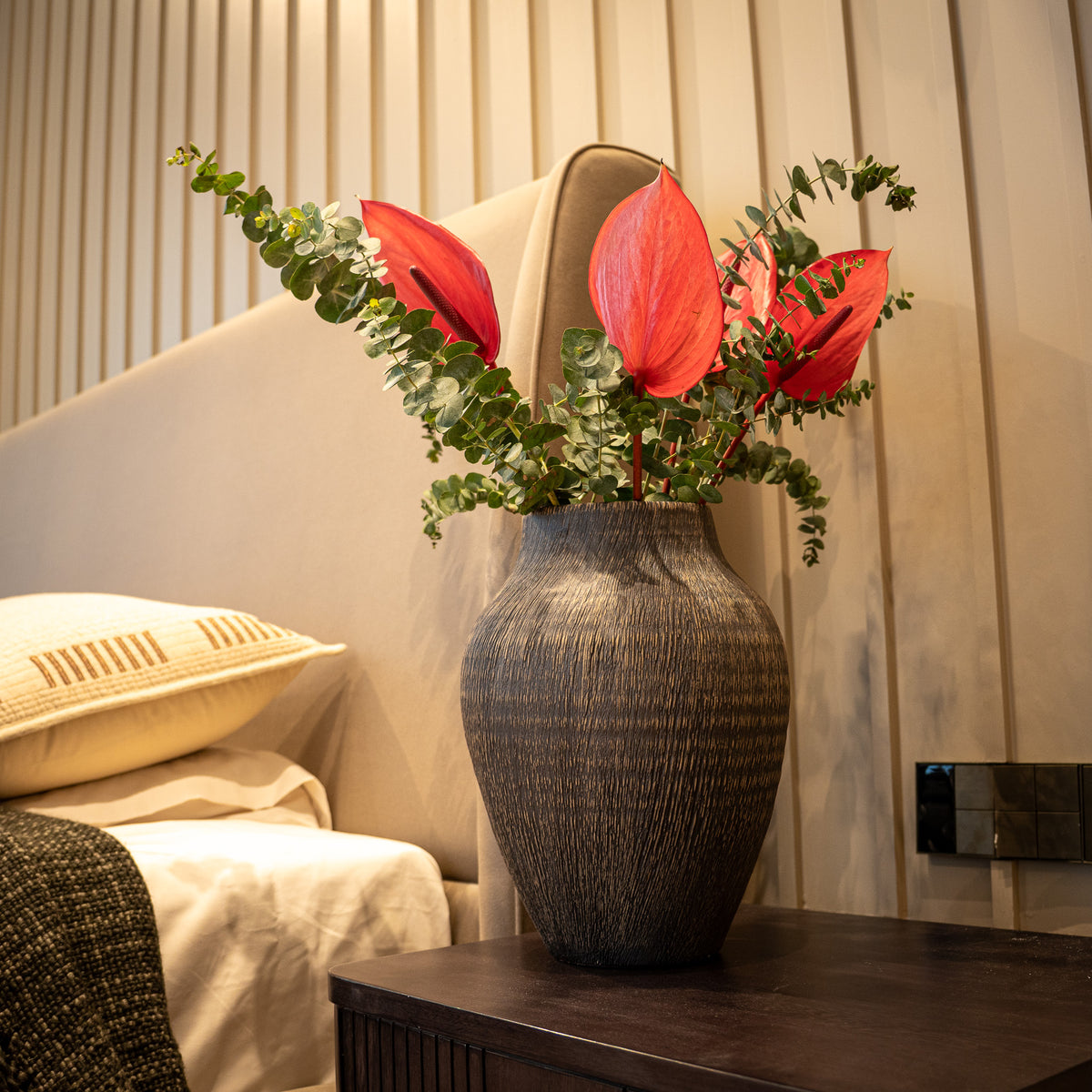 A dark textured ceramic vase filled with red anthuriums and green eucalyptus stems, placed on a dark wooden bedside table in a modern bedroom setting.