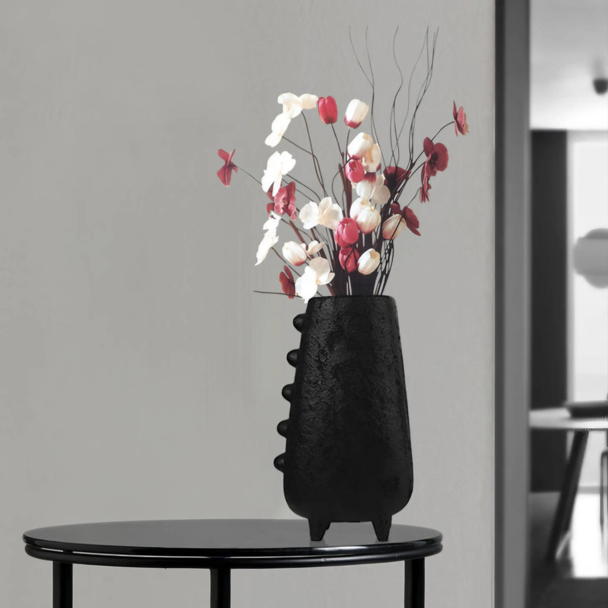 Black textured vase with flowers on a round black table against a gray wall.