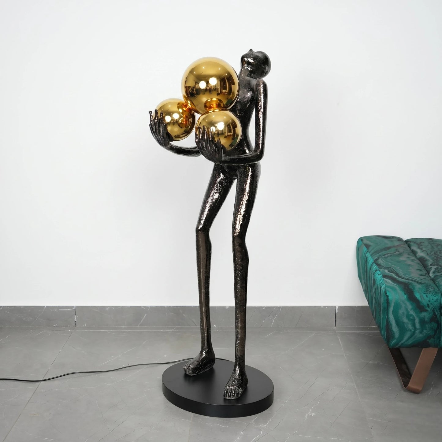 Decorative sculpture of a figure holding gold spheres on a gray floor with a white wall background.