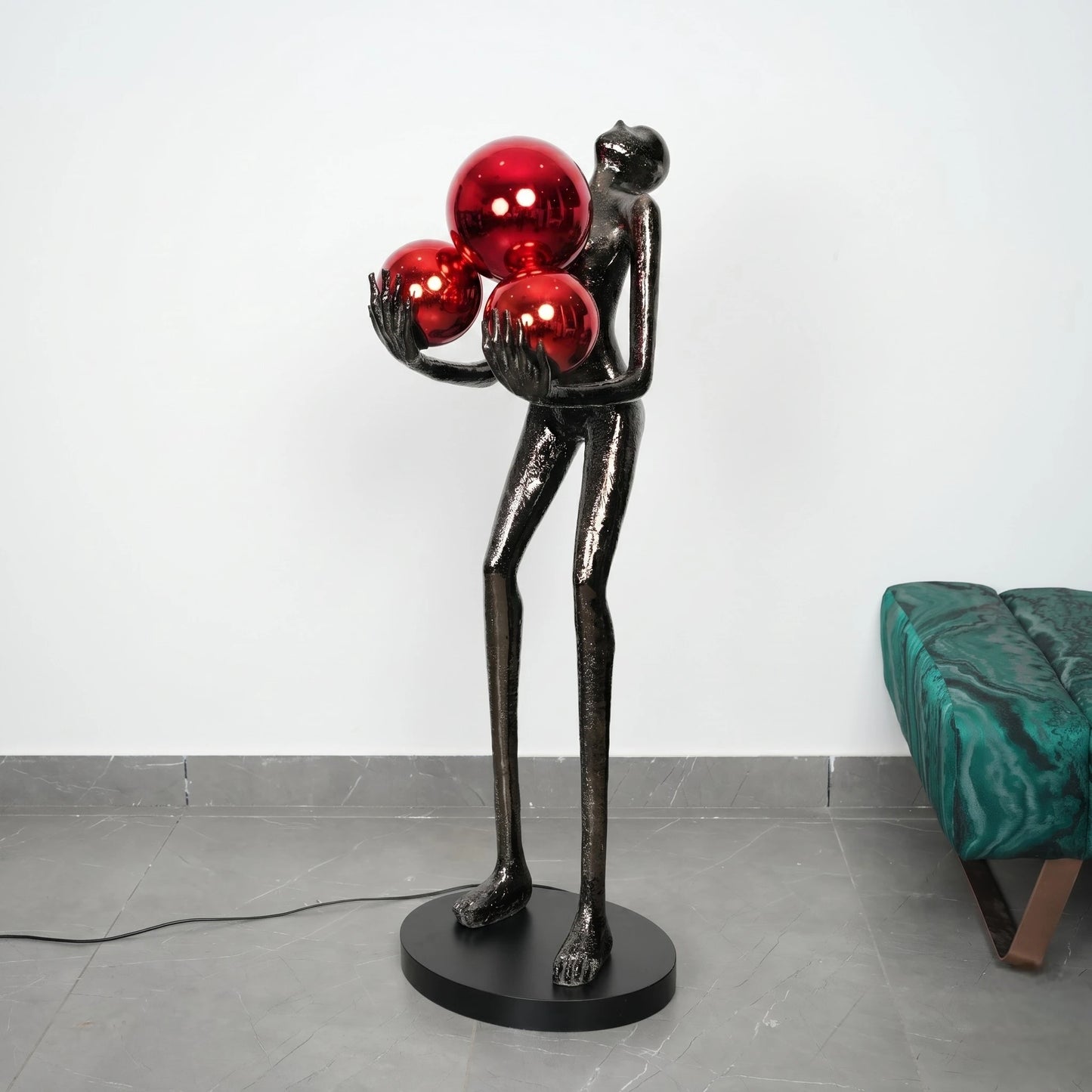 Statue of a figure holding three red spheres on a gray floor with a white wall background.