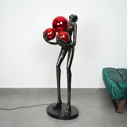 Statue of a figure holding three red spheres on a gray floor with a white wall background.